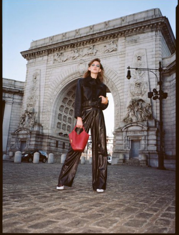 A model in black leather-like attire stands in front of a grand stone archway, holding a small red purse on a cobblestone street.