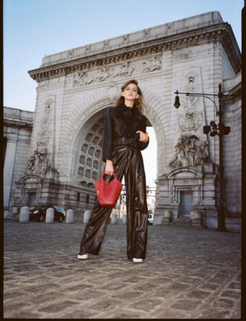 A model in black leather-like attire stands in front of a grand stone archway, holding a small red purse on a cobblestone street.