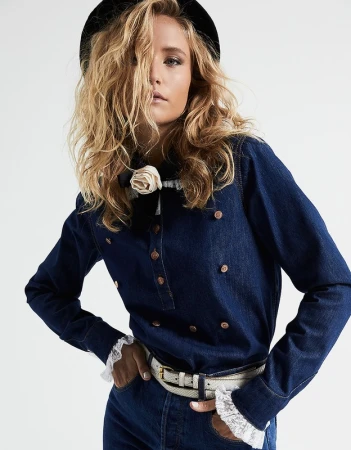 Blonde woman wearing a double breasted denim shirt, black hat and white belt against a white backdrop
