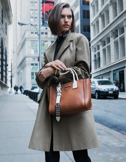 Stylish woman in trench coat holding designer handbag on city street. Brown leather purse with white accents and hardware.