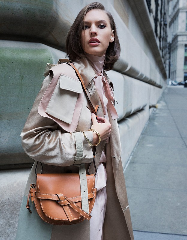 Attractive woman in a tan trench coat and blush dress stands in a city wearing a brown leather purse with concrete background.