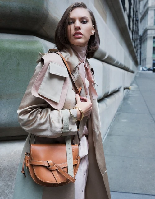 Attractive woman in a tan trench coat and blush dress stands in a city wearing a brown leather purse with concrete background.
