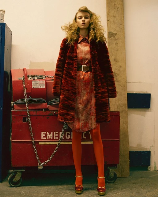 A young woman with curly hair poses in a vintage-style red coat, dress, tights, and heels. An emergency box is behind her.