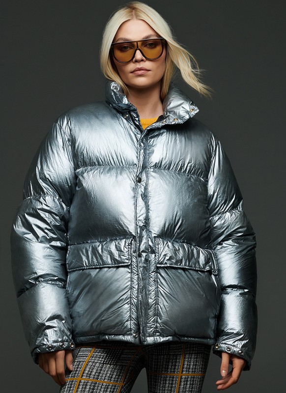 Woman poses in a shiny silver puffer jacket and sunglasses, with a gray backdrop and a glimpse of plaid pants at the bottom.