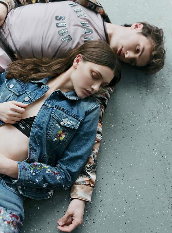 A fashionable couple reclines on a concrete floor. The female model is wearing a denim jacket with paint spatter, while the male is wearing an printed shirt.