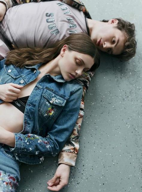A fashionable couple reclines on a concrete floor. The female model is wearing a denim jacket with paint spatter, while the male is wearing an printed shirt.