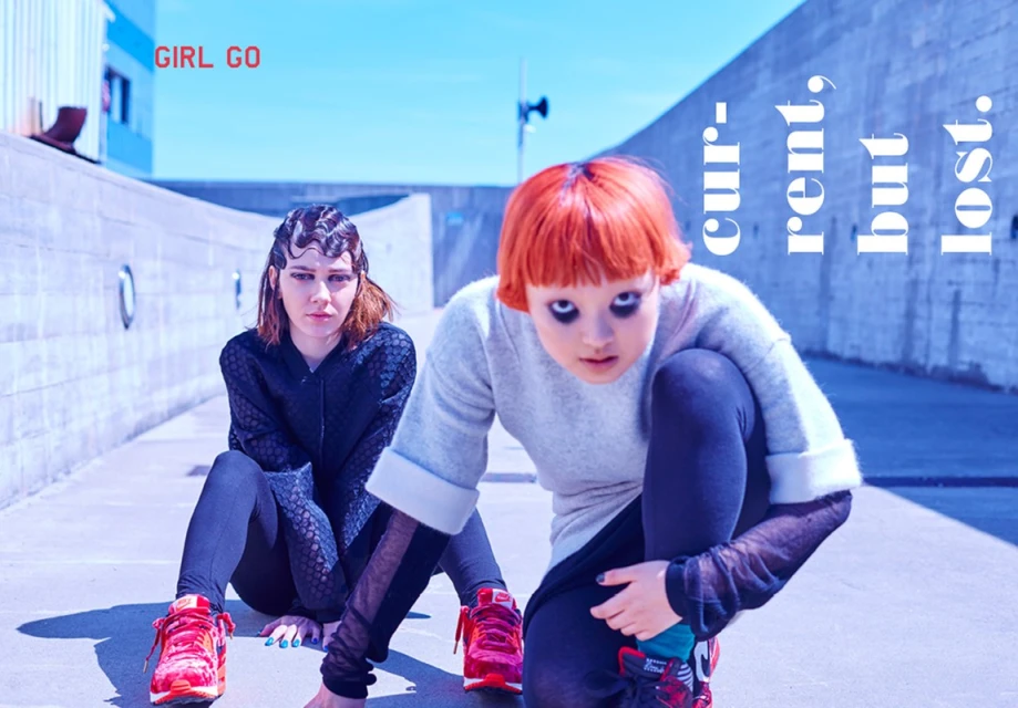 Two young women pose outdoors with vivid makeup and hair. One wears a textured jacket, the other a grey sweater, both with matching sneakers.