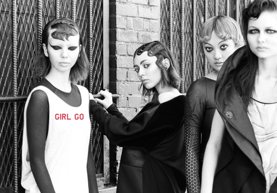 Black and white fashion photo of five young women in stylish outfits, posed against an urban backdrop of metal gates and brick walls.