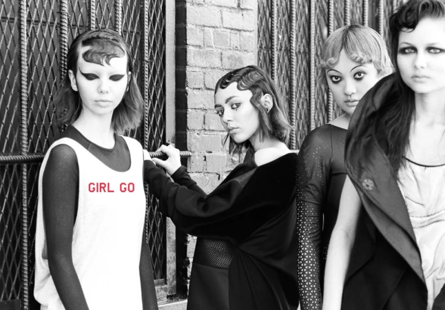 Black and white fashion photo of five young women in stylish outfits, posed against an urban backdrop of metal gates and brick walls.