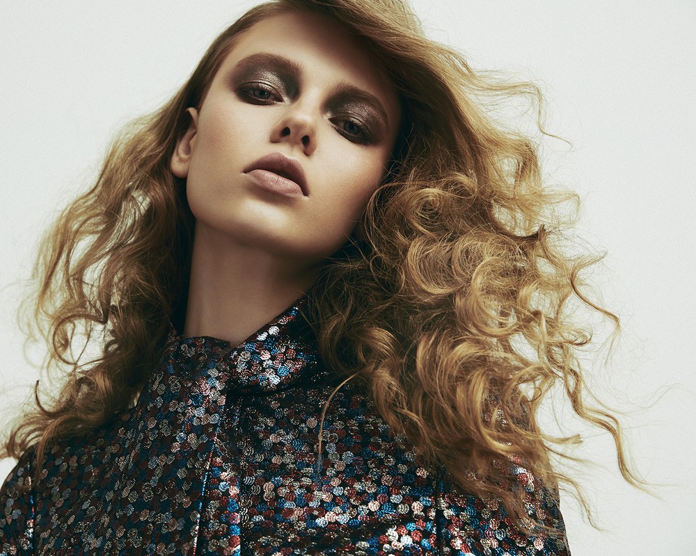 Close-up portrait of a fair-skinned adult woman with curly blonde hair, wearing dramatic eye makeup and a sequined blouse.