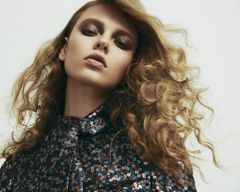Close-up portrait of a fair-skinned adult woman with curly blonde hair, wearing dramatic eye makeup and a sequined blouse.