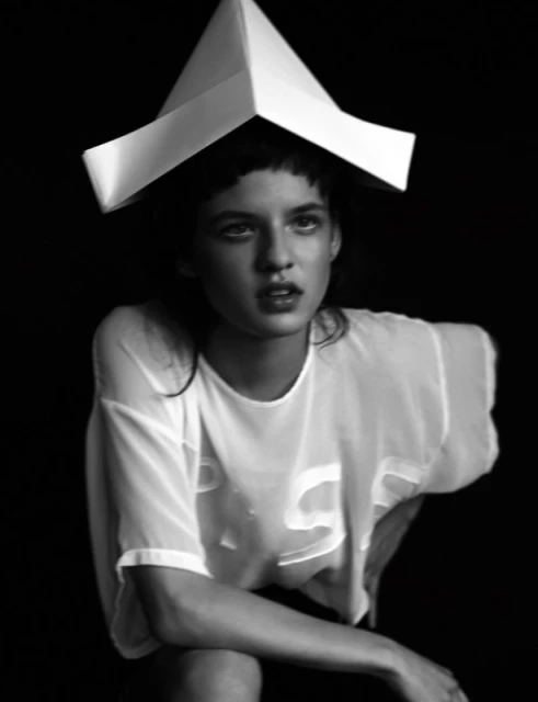 Portrait of a young woman wearing a paper hat and sheer shirt in a photo studio with black background.