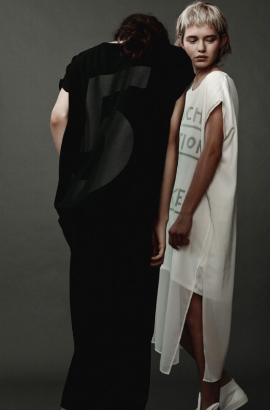 Two fashion models pose in a photo studio. One wears a black dress with a large number '5' on the back, the other a white dress with writing.