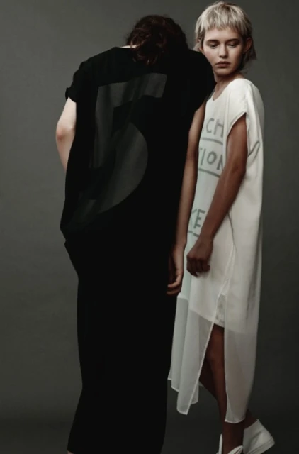 Two fashion models pose in a photo studio. One wears a black dress with a large number '5' on the back, the other a white dress with writing.