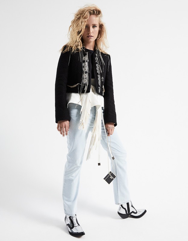 Full-length studio portrait of a blond woman in jeans and a black jacket with a white fringed belt and black and white boots, posing against a white backdrop.