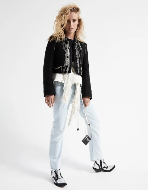 Full-length studio portrait of a blond woman in jeans and a black jacket with a white fringed belt and black and white boots, posing against a white backdrop.