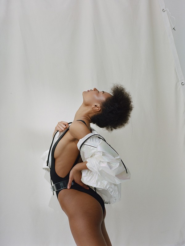 Portrait of a slender black woman with an afro, wearing a black bodysuit and clear plastic jacket, looking upwards in a light-colored setting.
