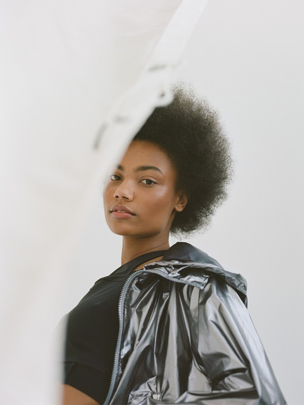 Headshot of a woman with afro wearing a black top and metallic jacket posing in front of a white backdrop.