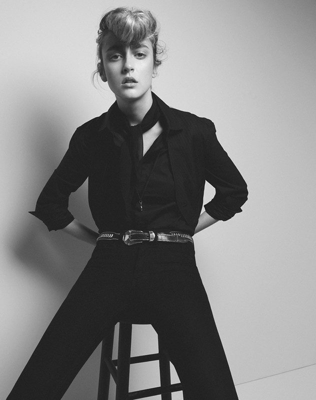 A young adult female model in a black blazer and pants is seated on a stool against a light background in a studio shot.