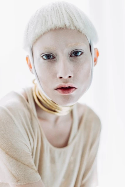 Close-up portrait of a person with fair skin, light hair, and a beige top wearing a layered golden necklace indoors against a blurred white backdrop.