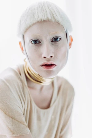 Close-up portrait of a person with fair skin, light hair, and a beige top wearing a layered golden necklace indoors against a blurred white backdrop.