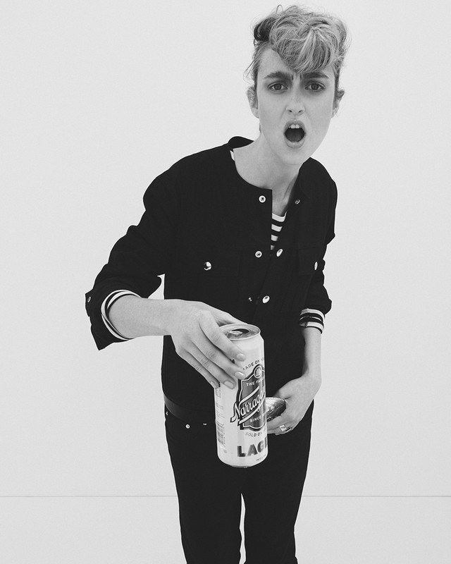Monochrome portrait of a young adult woman holding up a can of beer against a white backdrop in a high-key studio setting.