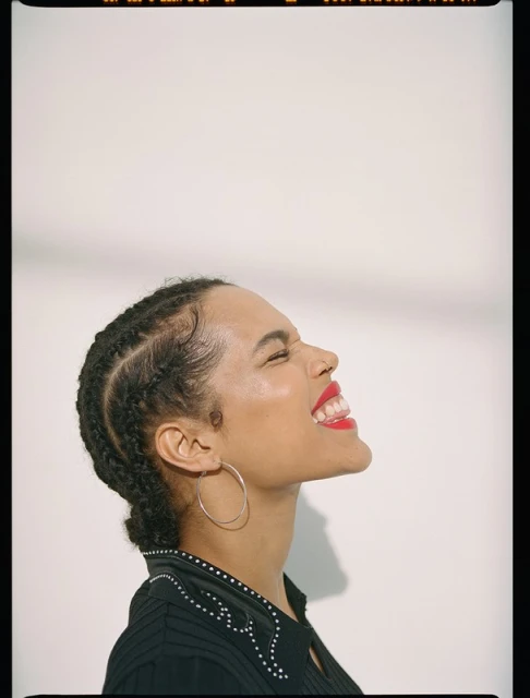Profile portrait of a woman with cornrow braids, red lipstick, and hoop earrings, expressing joy with closed eyes in a bright, neutral setting.