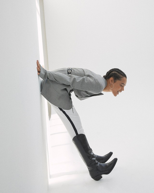 A Black woman in a gray jacket, white pants, and black boots presses against a white wall, smiling in a fashion-forward pose.
