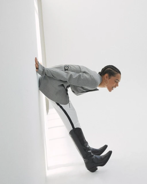 A Black woman in a gray jacket, white pants, and black boots presses against a white wall, smiling in a fashion-forward pose.