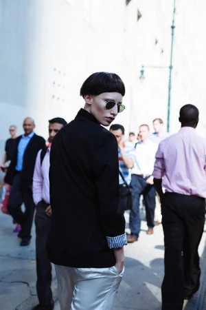 Adult female in sunglasses and a black blazer with silver pants posing in the street with a crowd of people in the background.