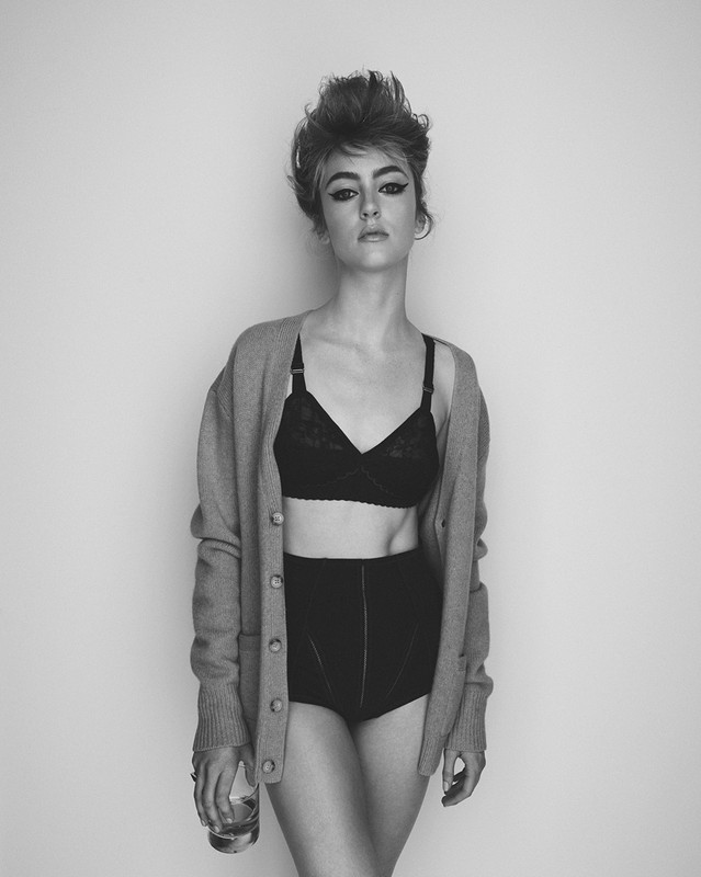 Black and white portrait of a young woman in lingerie and cardigan holding a glass, set against a plain light gray wall.