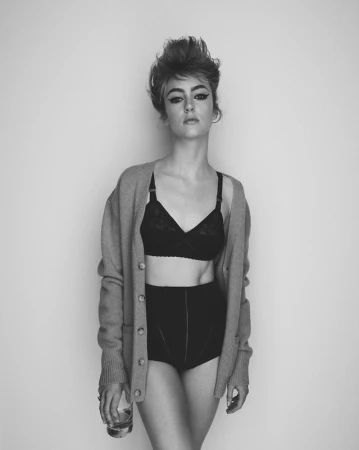 Black and white portrait of a young woman in lingerie and cardigan holding a glass, set against a plain light gray wall.
