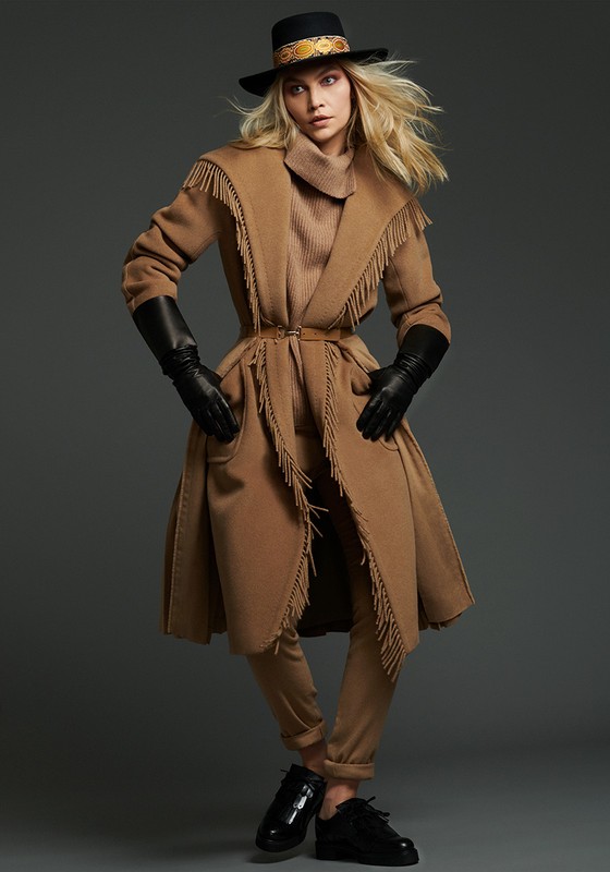 A woman with blonde hair posing in a tan coat with fringe, hat, black gloves and shoes against a grey studio backdrop.