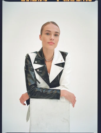 Woman in a black and white leather jacket leaning on a white block against a plain white background