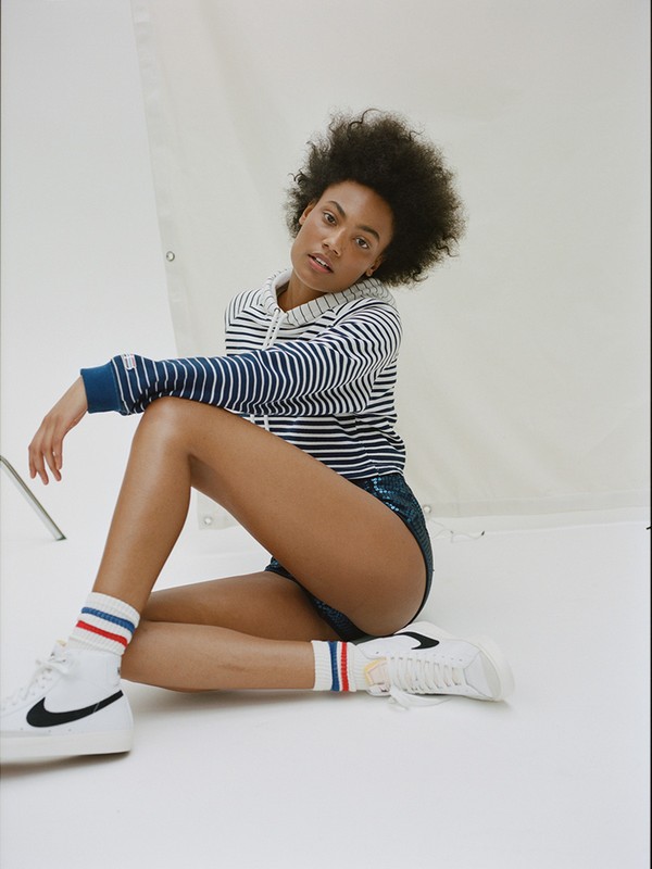 A woman with dark hair and striped top poses indoors with white sneakers and socks in a studio setting, gazing at the camera.