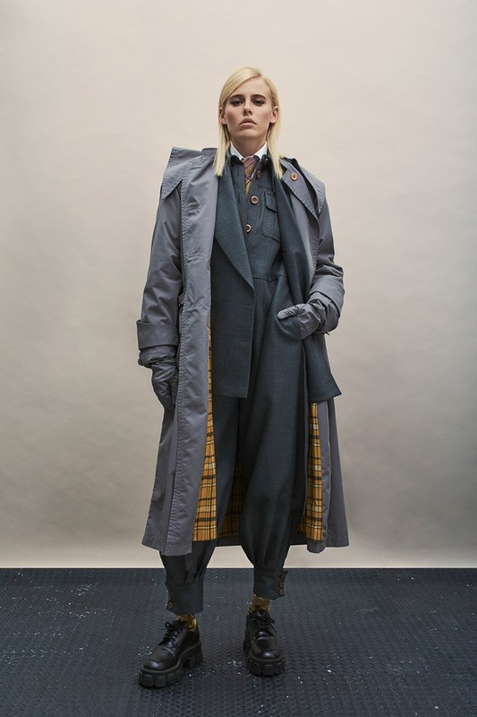A blonde adult posing in a grey trench coat over a suit in a studio, set against a neutral background, and styled for a fashion editorial.