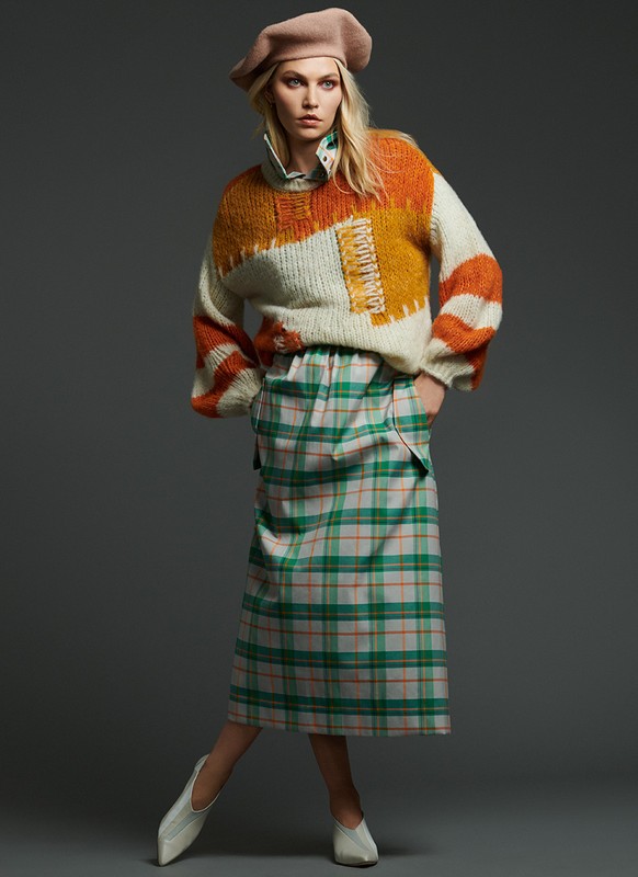 Adult fashion model in a studio portrait wearing a beige beret, cream-colored patterned sweater and plaid skirt on a grey background.