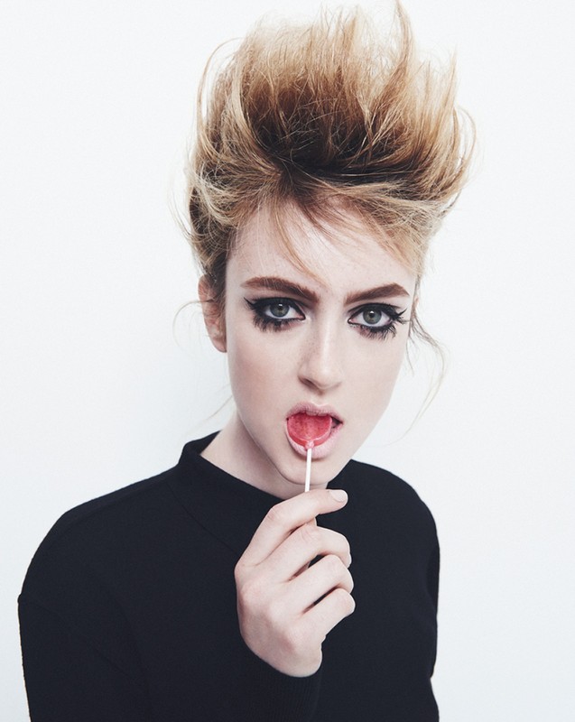 A fair-skinned woman with voluminous blonde hair and dramatic dark makeup is holding a lollipop in front of a plain white backdrop.