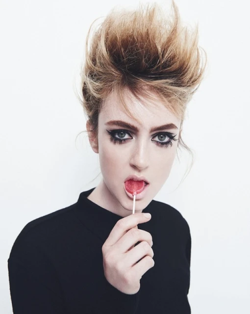 A fair-skinned woman with voluminous blonde hair and dramatic dark makeup is holding a lollipop in front of a plain white backdrop.