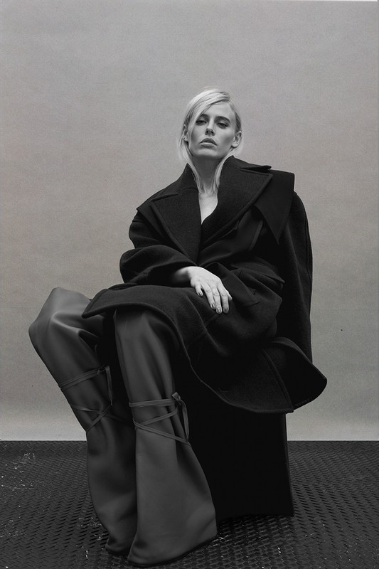 A solitary woman with blonde hair poses in a black coat and wide-legged trousers against a neutral backdrop in a studio setting.