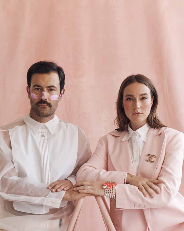 Portrait of a stylish couple posing in front of a pink background, wearing a light pink blazer and a semi-sheer shirt.
