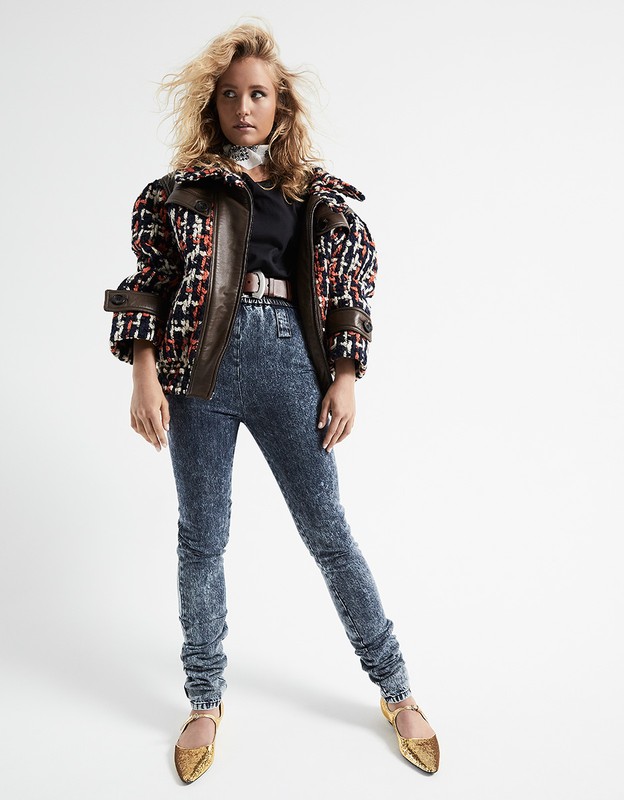Full studio shot of a woman with blonde hair wearing a patterned jacket, top, and blue leggings, with gold shoes posing on a white background.