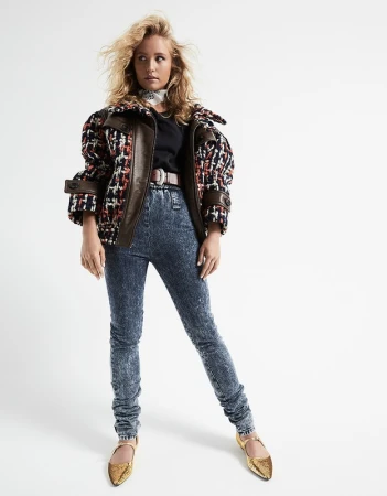 Model with tousled blonde hair wearing patterned tweed bomber jacket, acid wash jeans, and gold shoes against white background