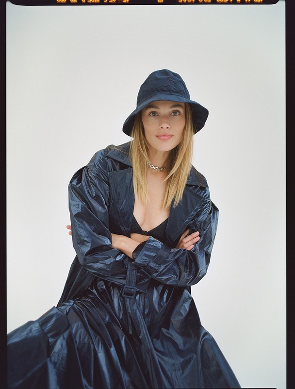 Model wearing a dark glossy trench coat and matching bucket hat against a plain light background