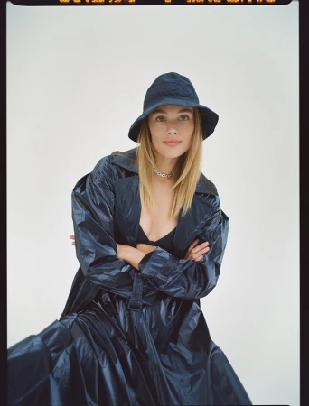 Model wearing a dark glossy trench coat and matching bucket hat against a plain light background