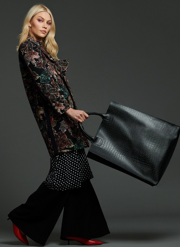 A woman strides in a floral coat, polka-dot skirt, and red shoes, carrying a large black handbag against a grey studio backdrop.