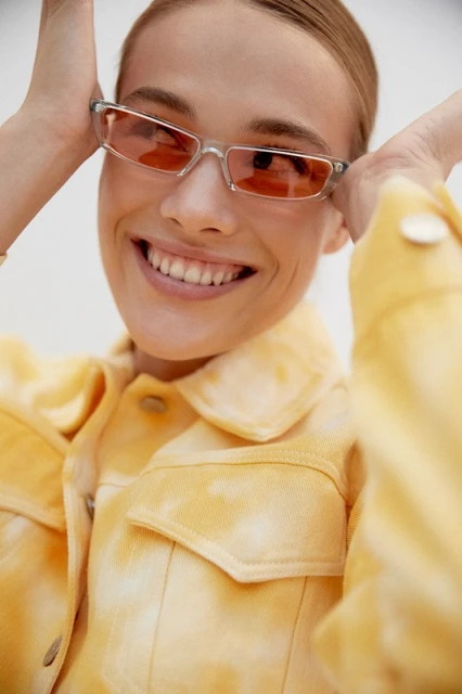 Smiling woman in a yellow tie-dye jacket and tinted glasses in a studio. Beauty shot against a white background with a cheerful facial expression.
