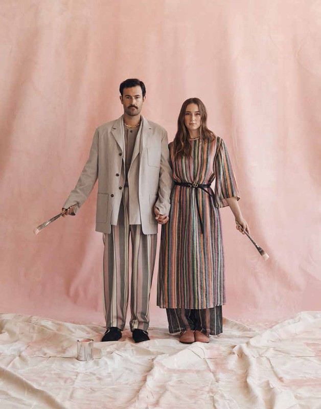 A stylish couple stands holding paintbrushes in a light-filled studio, wearing matching striped outfits, in an editorial fashion shoot.
