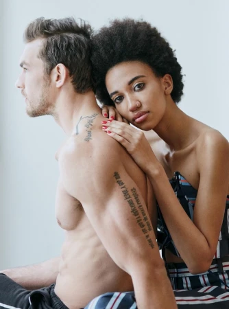 Portrait of a man with tattoos and a woman resting her head on his shoulder against a light background.