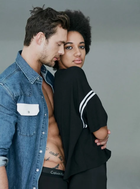 An intimate studio shot showcases an adult mixed-ethnicity couple, highlighting a bare-chested man in denim and a woman in a black top.